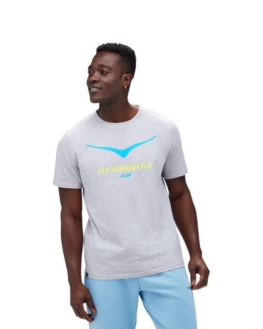Skön grå T-Shirt med Hoka loggan i blått på bröstkorgen. Under loggan står det "FLY HUMAN FLY" i grön färg.